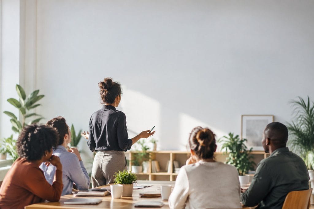 Une femme présente une idée devant un groupe diversifié de collègues assis autour d'une table dans un bureau lumineux.