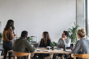 Une femme debout présente un projet à une équipe attentive assise autour d'une table dans un bureau moderne et lumineux.
